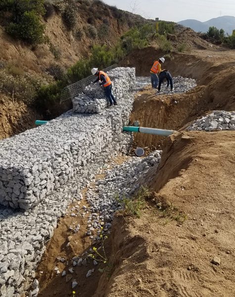 Trabajadores de CASFAM construyendo un muro de gaviones con malla metálica y piedra sobre una zanja con tubería expuesta, como parte de un proyecto de infraestructura civil en zona montañosa.
