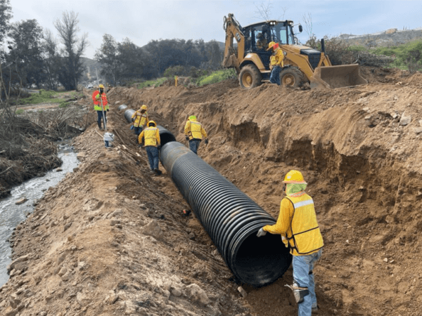 Equipo de trabajadores colocando un tubo corrugado negro en una zanja junto a una corriente de agua, mientras una retroexcavadora se posiciona detrás.