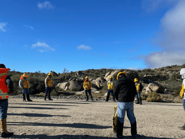 Grupo de trabajadores con chalecos reflejantes reunidos en círculo sobre un camino de tierra en una zona montañosa, realizando una junta de seguridad matutina.