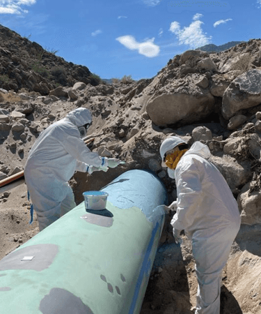 Dos trabajadores con overoles blancos y mascarillas aplican recubrimiento protector azul sobre una tubería verde en un terreno rocoso bajo un cielo parcialmente nublado.