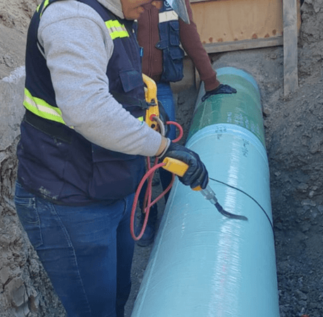 Trabajador con casco blanco y chaleco con reflejante inspecciona con un detector de poros una tubería grande de color verde menta en una zanja. Otro trabajador al fondo también revisa la superficie del ducto, dentro de un entorno de excavación.