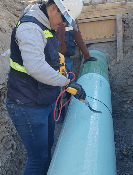 Trabajador con casco blanco y chaleco con reflejante inspecciona con un detector de poros una tubería grande de color verde menta en una zanja. Otro trabajador al fondo también revisa la superficie del ducto, dentro de un entorno de excavación.