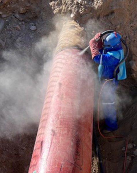 Trabajador con traje de protección azul y casco con respirador realiza proceso de sandblasting sobre un ducto rojo en una zanja, generando una nube de polvo a su alrededor. La actividad ocurre en un entorno de excavación con tierra suelta.