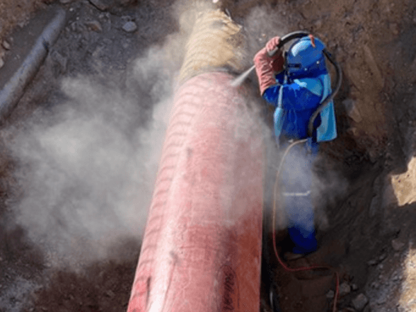 Trabajador con traje de protección azul y casco con respirador realiza proceso de sandblasting sobre un ducto rojo en una zanja, generando una nube de polvo a su alrededor. La actividad ocurre en un entorno de excavación con tierra suelta.