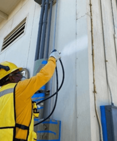 Trabajador con casco, careta y uniforme amarillo aplica pintura blanca con pistola pulverizadora en la esquina de un edificio industrial, junto a tubos eléctricos y cajas de control montadas en la pared. La acción parece formar parte de un mantenimiento o rehabilitación de infraestructura.