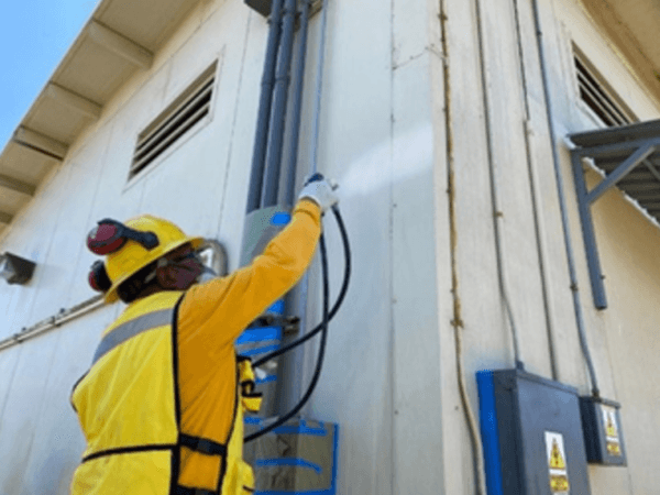 Trabajador con casco, careta y uniforme amarillo aplica pintura blanca con pistola pulverizadora en la esquina de un edificio industrial, junto a tubos eléctricos y cajas de control montadas en la pared. La acción parece formar parte de un mantenimiento o rehabilitación de infraestructura.