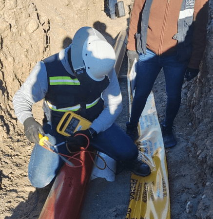 Trabajador con equipo de seguridad realiza una medición eléctrica sobre una tubería roja en una zanja, mientras una compañera supervisa junto a una cinta amarilla de advertencia para gas natural. Al fondo, se observa otro técnico con chaleco y casco blanco comunicándose por radio.