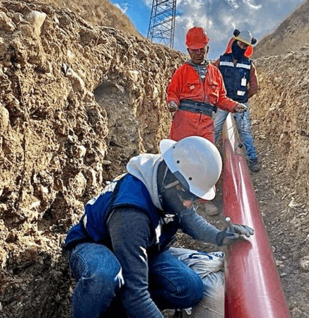 Trabajadores con equipo de protección inspeccionan y preparan una tubería roja colocada en una zanja profunda, rodeada de laderas de tierra. Uno de ellos realiza labores detalladas sobre la superficie del tubo, mientras otros supervisan el tramo alineado bajo una torre eléctrica.