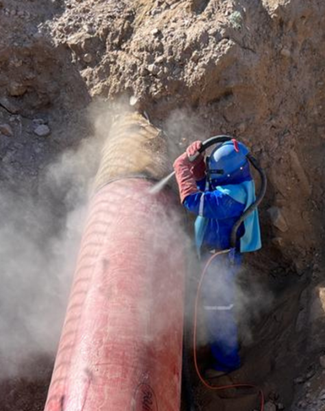 Trabajador con traje protector y casco realizando sandblasting sobre una tubería roja parcialmente enterrada. El proceso genera una nube de polvo, indicando limpieza abrasiva en un entorno de excavación.