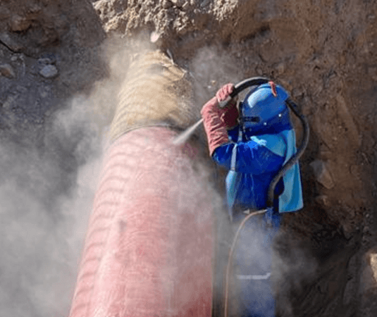 Trabajador con traje protector y casco realizando sandblasting sobre una tubería roja parcialmente enterrada. El proceso genera una nube de polvo, indicando limpieza abrasiva en un entorno de excavación.