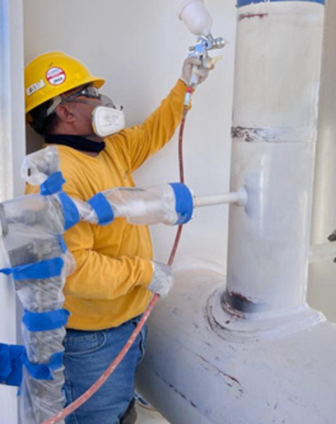 Trabajador con casco y mascarilla aplicando pintura blanca con pistola de aire sobre tubería industrial. Las válvulas y uniones están protegidas con plástico y cinta azul, en una instalación de gas natural al aire libre.