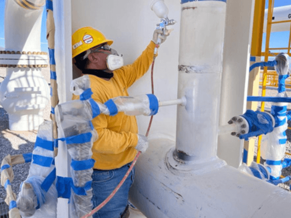 Trabajador con casco y mascarilla aplicando pintura blanca con pistola de aire sobre tubería industrial. Las válvulas y uniones están protegidas con plástico y cinta azul, en una instalación de gas natural al aire libre.