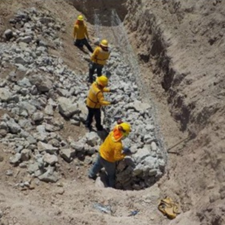 Cuatro trabajadores con uniforme amarillo y casco construyendo un muro de gaviones dentro de una zanja, rodeados de tierra y piedras grandes. Están colocando las rocas dentro de una malla metálica.