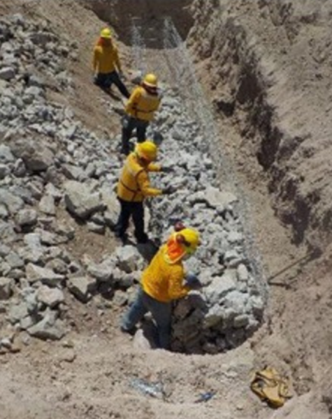 Cuatro trabajadores con uniforme amarillo y casco construyendo un muro de gaviones dentro de una zanja, rodeados de tierra y piedras grandes. Están colocando las rocas dentro de una malla metálica.