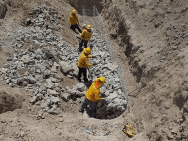 Cuatro trabajadores con uniforme amarillo y casco construyendo un muro de gaviones dentro de una zanja, rodeados de tierra y piedras grandes. Están colocando las rocas dentro de una malla metálica.