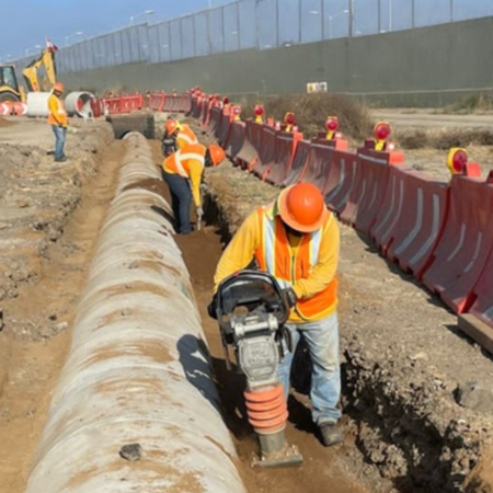 Trabajadores con chalecos y cascos naranjas realizando labores de compactación de tierra junto a una zanja con tubería de concreto, en una zona de construcción delimitada por barreras de seguridad rojas y grises.