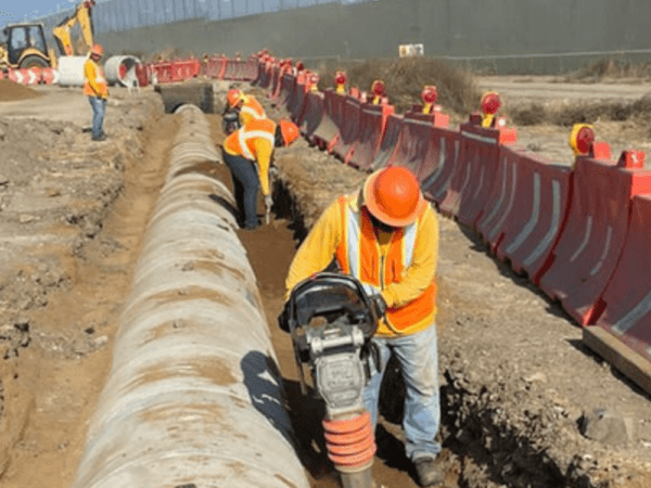 Trabajadores con chalecos y cascos naranjas realizando labores de compactación de tierra junto a una zanja con tubería de concreto, en una zona de construcción delimitada por barreras de seguridad rojas y grises.
