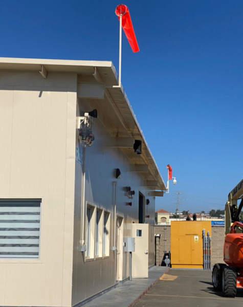 Dos trabajadores con arnés de seguridad en una plataforma elevadora tipo brazo articulado, pintando la parte superior de un edificio beige con ventanas, bajo un cielo azul y una manga de viento roja ondeando en el techo.