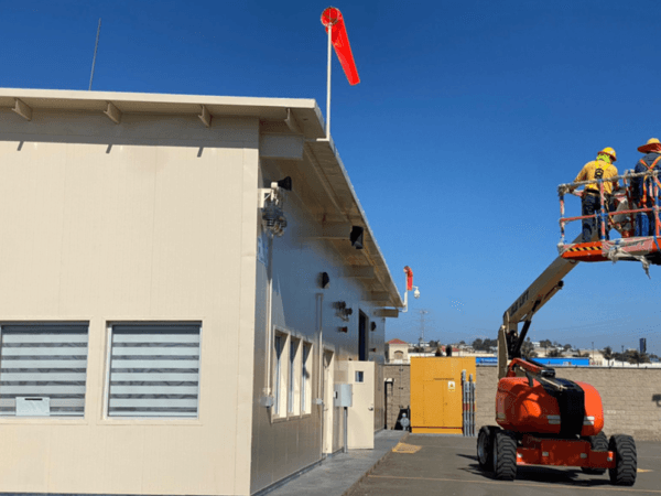 Dos trabajadores con arnés de seguridad en una plataforma elevadora tipo brazo articulado, pintando la parte superior de un edificio beige con ventanas, bajo un cielo azul y una manga de viento roja ondeando en el techo.