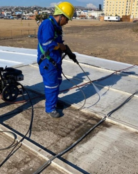 Trabajadores con equipo de protección pintando la fachada de un edificio industrial beige, usando escaleras, brochas y rodillos, bajo cielo despejado y junto a una manga de viento en la esquina del techo.