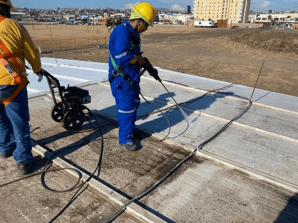 Trabajadores con equipo de protección pintando la fachada de un edificio industrial beige, usando escaleras, brochas y rodillos, bajo cielo despejado y junto a una manga de viento en la esquina del techo.