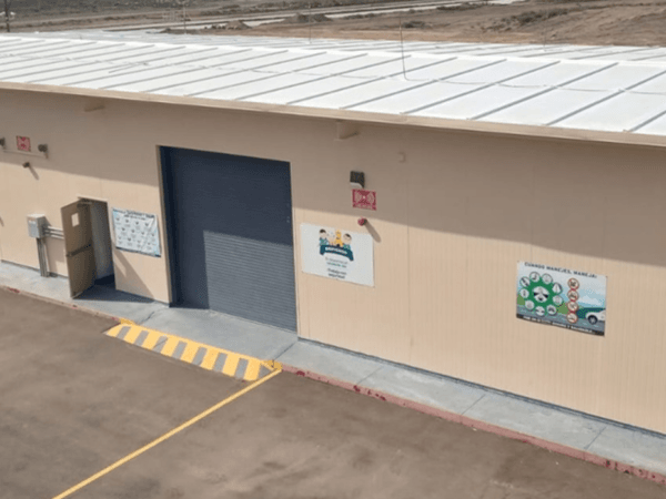 Edificio industrial beige con techo blanco visto desde un ángulo elevado, con rampas de acceso, señalamientos de seguridad, manga de viento roja y espacio de estacionamiento amplio sobre asfalto.