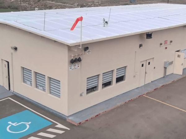 Vista aérea de un edificio industrial beige con techo blanco, estacionamiento pavimentado, espacio reservado para personas con discapacidad y manga de viento en el techo, todo ubicado en un entorno de obra.