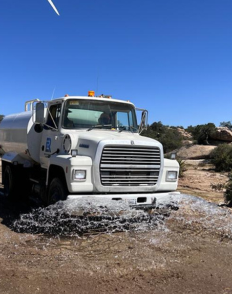 Camión cisterna blanco esparciendo agua sobre camino de terracería durante jornada de trabajo, con fondo de aerogenerador y paisaje rocoso y desértico bajo cielo despejado.