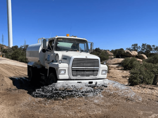 Camión cisterna blanco esparciendo agua sobre camino de terracería durante jornada de trabajo, con fondo de aerogenerador y paisaje rocoso y desértico bajo cielo despejado.
