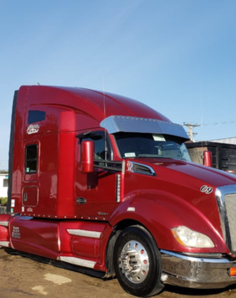 Tracto camión Kenworth color rojo cereza estacionado sobre un terreno de tierra, con luz solar brillante y fondo de maquinaria y remolques industriales.