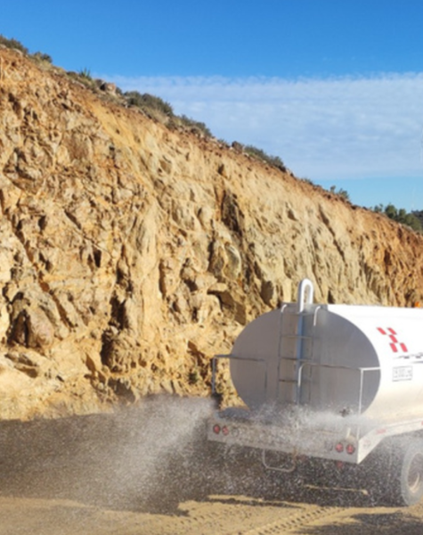 Camión cisterna blanco esparciendo agua sobre un camino de terracería, con un acantilado rocoso a un lado y un aerogenerador visible al fondo bajo un cielo despejado.