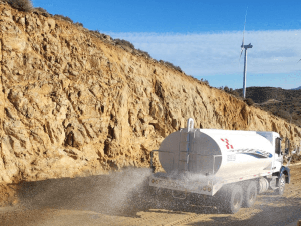 Camión cisterna blanco esparciendo agua sobre un camino de terracería, con un acantilado rocoso a un lado y un aerogenerador visible al fondo bajo un cielo despejado.
