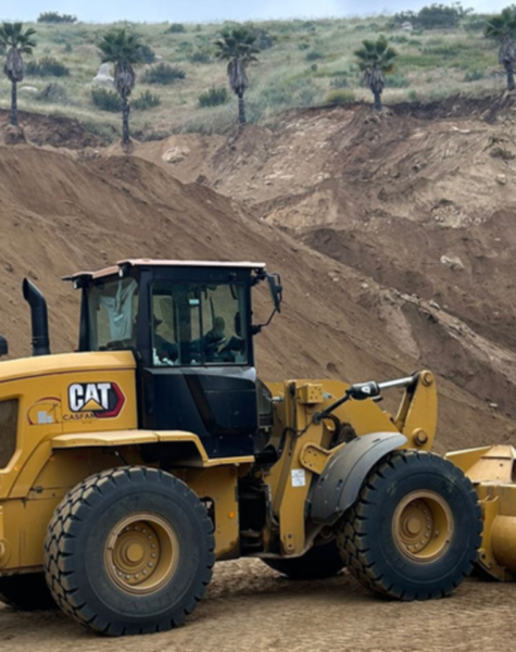 Cargadora frontal CAT 930K con logotipo de CASFAM en operación sobre terreno terroso, con un fondo de ladera y palmas dispersas en la parte superior del paisaje.