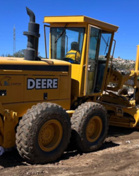 Motoniveladora John Deere operada por personal de CASFAM trabajando en el aplanado de un camino de terracería, con montañas al fondo y un aerogenerador visible en el paisaje.