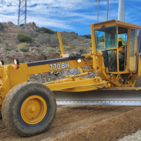 Motoniveladora John Deere 770 BH en operación sobre un camino de terracería, nivelando terreno en una zona rural con colinas y postes eléctricos al fondo.