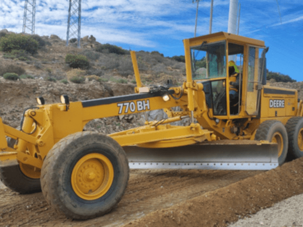 Motoniveladora John Deere 770 BH en operación sobre un camino de terracería, nivelando terreno en una zona rural con colinas y postes eléctricos al fondo.