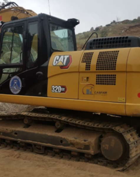 Excavadora CAT modelo 320 GX operando en una zona rocosa con terreno de tierra, mostrando logotipos de seguridad, la marca CASFAM y distintivos de operación pesada.