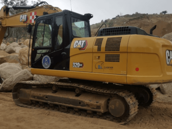 Excavadora CAT modelo 320 GX operando en una zona rocosa con terreno de tierra, mostrando logotipos de seguridad, la marca CASFAM y distintivos de operación pesada.